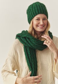 Set de căciulă și eșarfă tricotate verzi, având un design cu rigle. Purtați peste un pulover crem, tricotat cu model de sfoară. Textură moale, aspect cald.