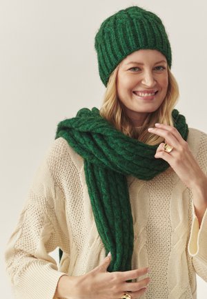 Set de căciulă și eșarfă tricotate verzi, având un design cu rigle. Purtați peste un pulover crem, tricotat cu model de sfoară. Textură moale, aspect cald.
