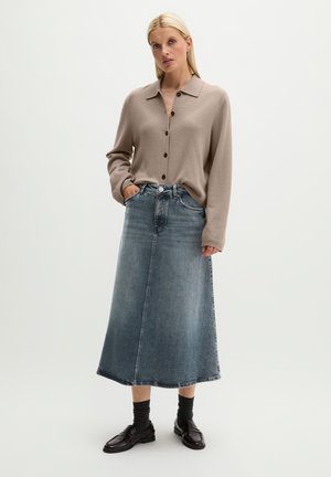 Femme portant un pull beige boutonné, une jupe midi en denim bleu, des chaussettes noires et des mocassins en cuir noir, debout devant un fond uni.