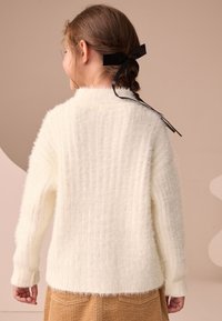 Pull en maille côtelée blanc avec une finition texturée et manches longues, orné d'un nœud à l'arrière. Associé à une jupe en velours côtelé.