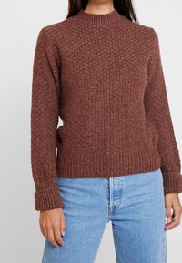 Femme portant un pull en maille texturée marron avec des poignets repliés, associé à un jean taille haute bleu clair, se tenant devant un fond uni.