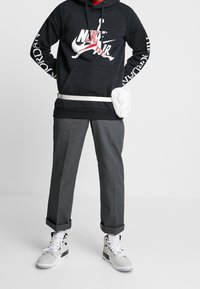 Felpa nera con cappuccio e logo grafico bianco; pantaloni grigi a gamba dritta; borsa a tracolla bianca; sneakers alte con motivo.