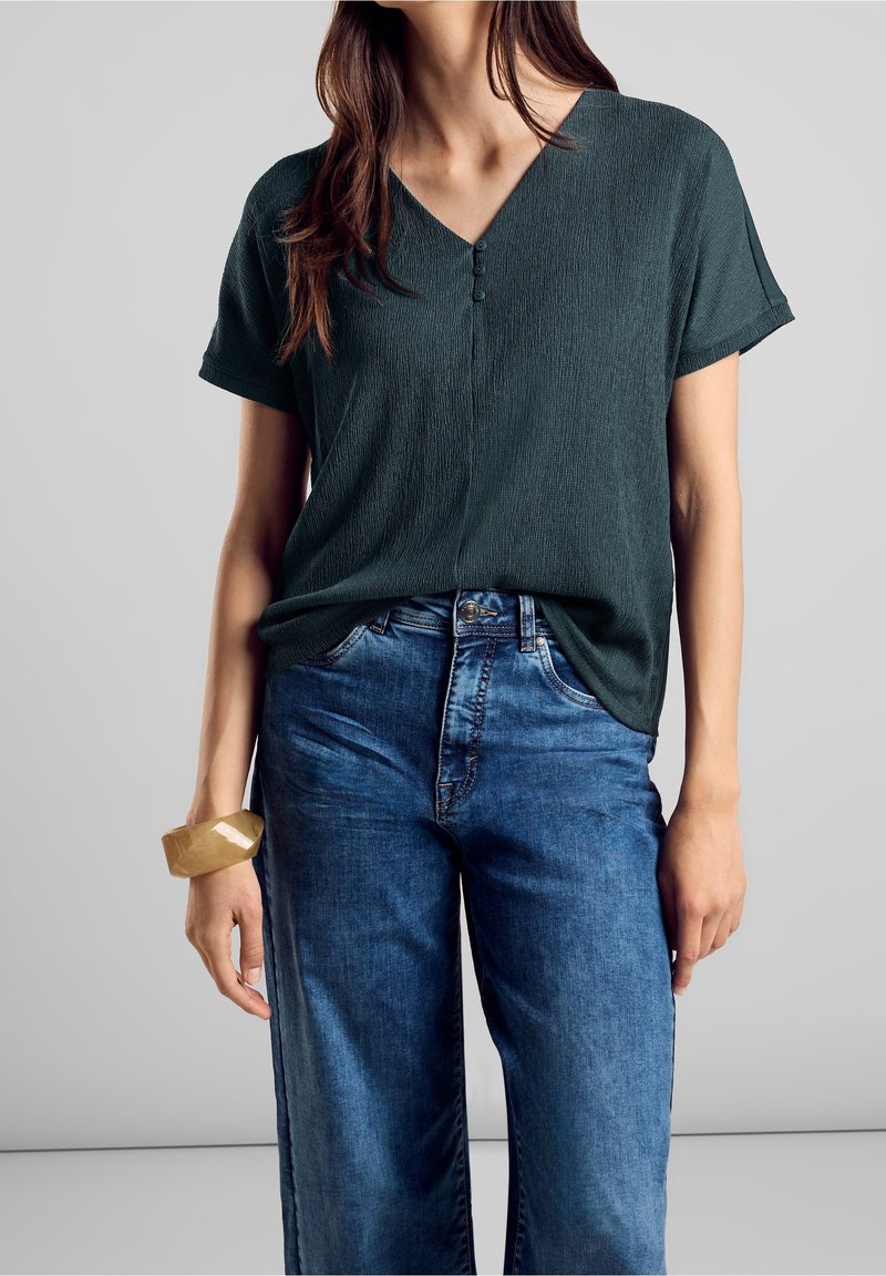 Haut en v-texture vert foncé à manches courtes et trois boutons à l'avant, associé à un jean large en denim bleu taille haute.