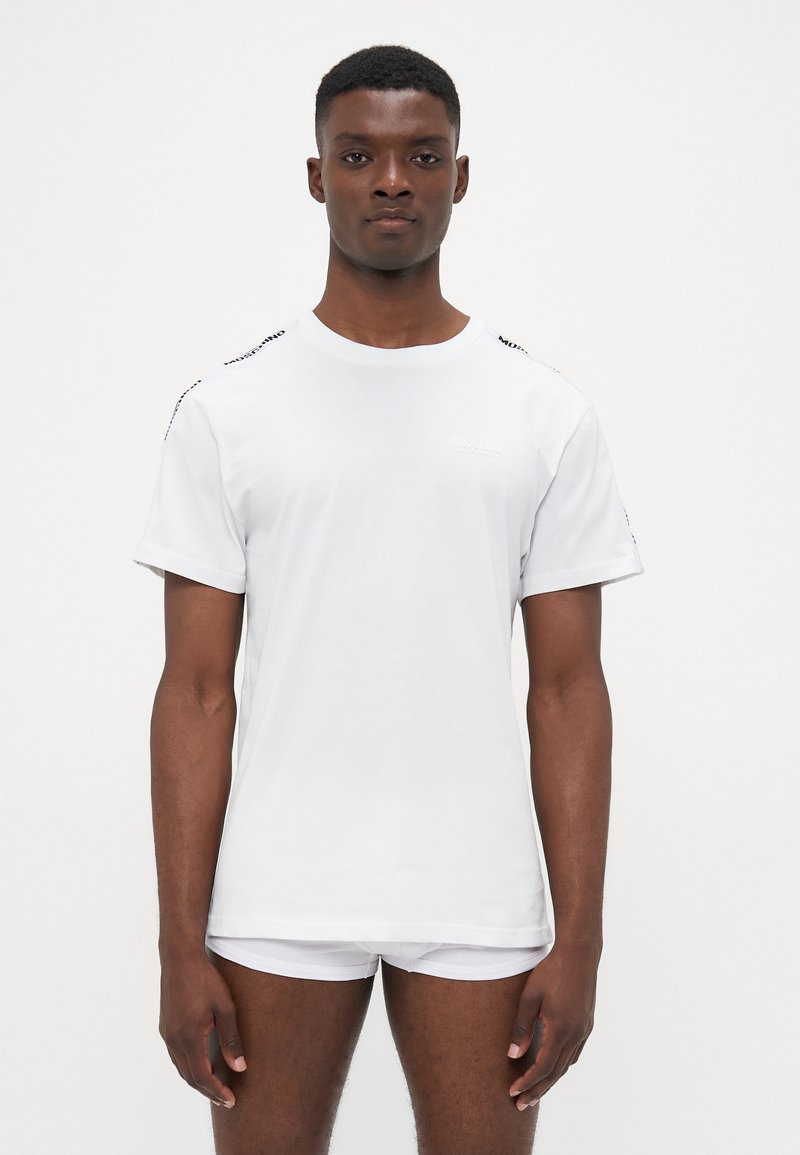 Vit t-shirt i bomull, med rund hals, korta ärmar och svarta logotypdetaljer på axlarna. Slät textur, standard passform.