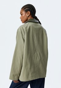 Olivgrön oversized jacka med en mjuk svart krage, texturerat tyg och avslappnad passform. Har en rak skärning och minimala sömnadsdetaljer.