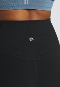 Magasan fazonú fekete leggings sima textúrával, fehér virágos logó részlettel a derékpánton és kontrasztos varrással.