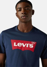 Hombre vistiendo una camiseta azul marino con un gran logo rojo y blanco de Levi's en el pecho, fondo neutro.