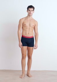 Boxeri de culoare albastru marin, cu talie elastică roșie, având logo-ul "Tommy Hilfiger". Modelul este fără cămașă, stând pe un fundal simplu.