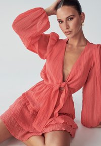 Frau mit slicked-back Haar, die ein korallenfarbenes, strukturiertes Kleid mit aufgepufften langen Ärmeln und vornem Bindeband trägt. Sie posiert vor einem schlichten, hellen Hintergrund.