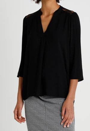 Femme portant un chemisier noir à col en V avec des détails en dentelle sur les épaules et des manches trois-quarts, assorti à une jupe pied-de-poule noir et blanc.