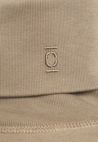 Beige värillinen ribbineuloksinen tekstiili, jossa on hienovarainen kirjailtu logo. Kangas on hienorakenteinen ja siinä on taitettu reuna, mikä osoittaa venyvyyttä ja kestävyyttä.