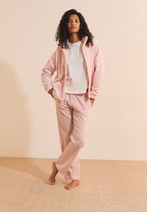Femme aux cheveux bouclés portant une veste polaire rose clair et un pantalon, debout pieds nus sur un sol beige devant un mur beige uni.
