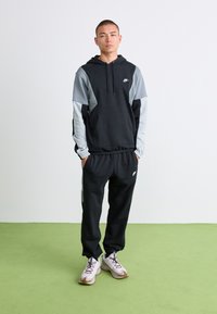 Young man standing with hands in pockets wearing black and grey Nike hoodie, matching black Nike joggers, and white sneakers on green floor.