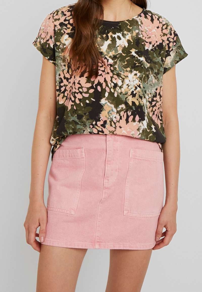 Femme portant une chemise à manches courtes à motif floral rose et vert, associée à une mini-jupe rose avec des poches plaquées à l'avant.
