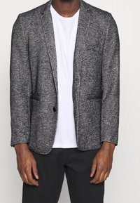 Grijze gebreide blazer met een notch-lapel, enkele knoopsluiting, twee voorzakken, gedragen over een eenvoudige witte t-shirt.