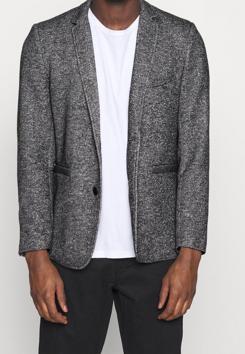 Grijze gebreide blazer met een notch-lapel, enkele knoopsluiting, twee voorzakken, gedragen over een eenvoudige witte t-shirt.