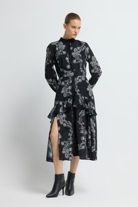 Schwarzes Kleid mit Paisley-Muster aus leichtem Stoff, mit langen Ärmeln, hohem Kragen, gestuften Rüschen und Seitenschlitzen. Kombiniert mit schwarzen Stiefeletten.