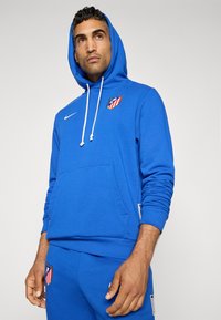 Blauer Kapuzenpullover aus weichem Stoff, mit einer Fronttasche, weißen Kordeln, einem Nike-Logo und dem Atlético Madrid-Wappen auf der Brust.