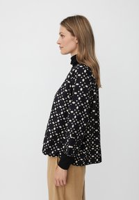 Schwarzes Oberteil mit weißem geometrischem Muster, hoher Ausschnitt, lockere Passform, lange Ärmel. Kombiniert mit beigen Hosen, die ein einfaches Design aufweisen.