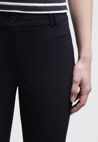 Pantaloni aderenti di colore blu navy scuro realizzati in tessuto liscio. Presentano una parte frontale piatta, tasche laterali e un'unica chiusura con bottone.