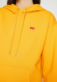 Sudadera amarilla de Levi's con bolsillo frontal, capucha con cordón y agujas plateadas. Pequeño logo rojo en la parte izquierda del pecho.