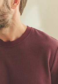 Burgundy kötött pulcsi texturált mintával, bordás kerek nyakkivágással és puha anyaggal, amely a laza dizájnt hangsúlyozza.