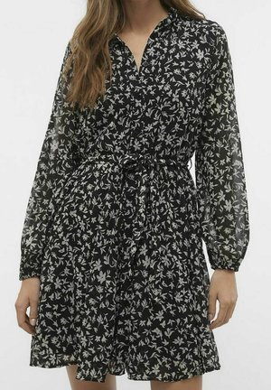 Femme portant une robe noire à imprimé floral blanc, manches longues semi-transparentes, devant boutonné et ceinture nouée à la taille.