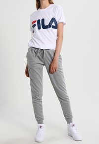 White t-shirt with large blue and red FILA logo, paired with gray sweatpants and white sneakers, featuring a soft, casual fit.