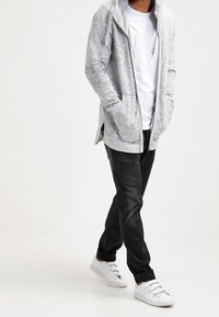 Sudadera con cremallera de color gris claro, con bolsillos laterales, llevada sobre una camiseta blanca. Unos pantalones vaqueros ajustados negros y unas zapatillas blancas completan el look.