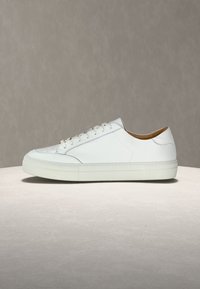 Λευκό δερμάτινο χαμηλό αθλητικό παπούτσι με λευκά κορδόνια και μπεζ εσωτερική επένδυση πάνω σε ανοιχτή πέτρινη επιφάνεια και γκρι φόντο.