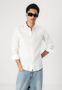Hvid skjorte med knaplukning i letvægtsstof, med lange ærmer rullet op ved manchetterne, parret med løstsiddende lyseblå jeans og mørke solbriller.