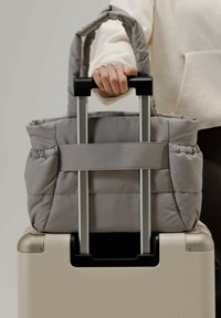 Borsa tote grigia imbottita con manici doppi fissati al manico telescopico di una valigia beige; presenta una tracolla piatta e un tessuto testurizzato.