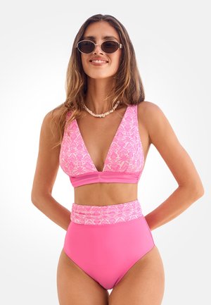 Kobieta w różowym bikini z wysokim stanem i kwiatowym wzorem na górze, z muszelkowym naszyjnikiem i owalnymi okularami przeciwsłonecznymi, uśmiechająca się z rękami za plecami.