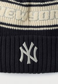 Črno-kremna pletena kapa z rebrasto teksturo, vodoravnimi črtami in izvezenim napisom "NEW YORK" ter logotipom Yankees v sivi barvi.