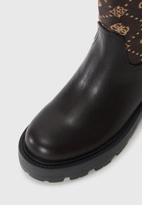 Bruine leren enkelboot met een ronde neus, gestructureerde rubberen zool en een bovenwerk van stoffen met een beige patroon op een donkere achtergrond.