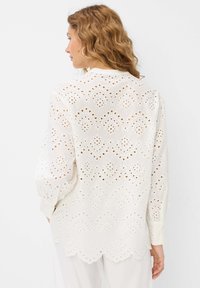 Weiße Bluse mit Lochstickerei, gewelltem Saum und langen Ärmeln. Sie verfügt über ein florales Muster mit ausgeschnittenen Details für Atmungsaktivität.