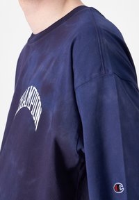 Sudadera azul marino con un logo blanco de "Champion" en el pecho y un pequeño logo bordado en la manga izquierda. Textura suave, cuello redondo.