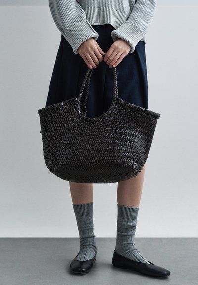 Πλεκτή μαύρη τσάντα tote με διπλές λαβές, διαθέτει υφή στην επιφάνεια και ευρύχωρο εσωτερικό, συνδυασμένη με σκουρόχρωμη φούστα και γκρι καλτσάκια μέχρι το γόνατο.