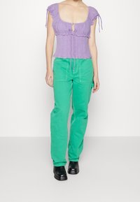 Top in lavanda con arricciature e maniche a volant, abbinato a pantaloni cargo verdi con tasche frontali e cuciture a contrasto. Stivali neri con tacco.