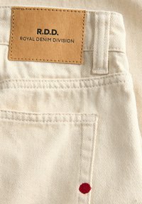 Tissu en jean beige clair avec poche arrière, passant de ceinture cousu et étiquette en cuir marron clair avec texte noir "R.D.D. Royal Denim Division".