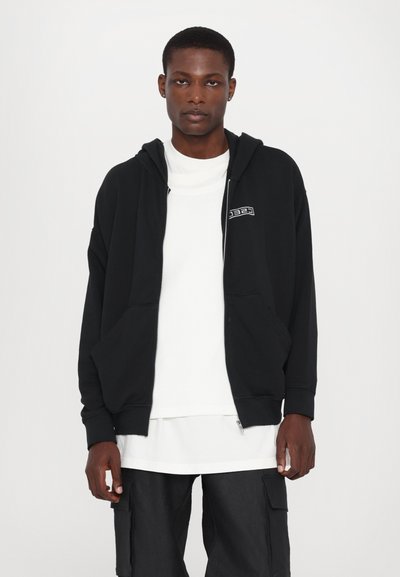 Black zip-up hoodie made of soft fabric, featuring a front pocket and logo patch. Worn over a white shirt and paired with black cargo pants.