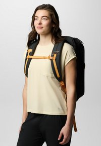 Schwarzer Rucksack mit orangefarbenen Akzenten, ausgestattet mit gepolsterten Trägern und einem Brustclip. Modell trägt ein hellgelbes Hemd und schwarze Hosen.