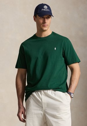 Homme portant un t-shirt polo vert foncé avec logo, un short blanc, une casquette Ralph Lauren marine et une montre avec un bracelet marine, posant avec une main dans la poche.