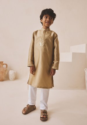 Ein junges Mädchen steht lächelnd da, trägt eine goldbestickte Kurta mit weißen Hosen und braunen Sandalen vor einem neutralen Hintergrund mit Vasen.