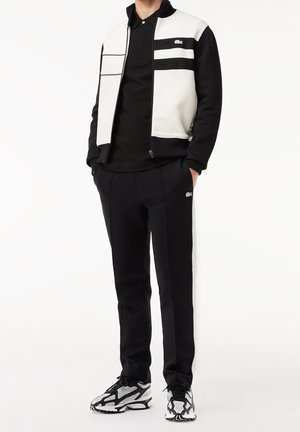 Veste zippée noire et blanche avec un devant texturé, polo noir en dessous, pantalons noirs avec des bandes blanches sur les côtés et baskets noires et argentées à motifs.