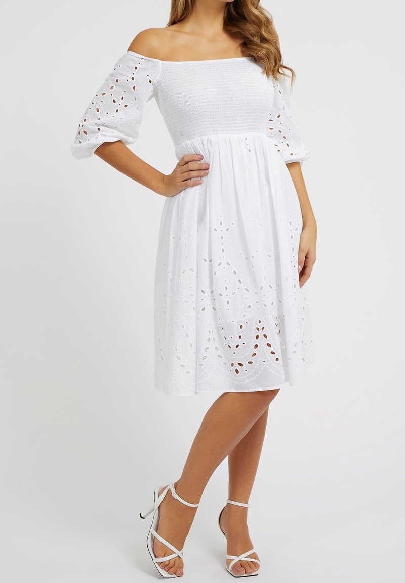 Femme portant une robe blanche à épaules dénudées avec des manches bouffantes et des détails en œillets, associée à des sandales blanches à talons aiguilles à brides.