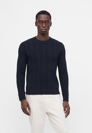 Homme portant un pull torsadé bleu foncé et un pantalon blanc, debout devant un fond clair uni.