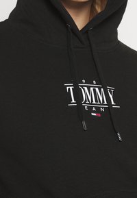 Černá mikina s kapucí a stahovacími šňůrkami, s bílým logem "1985 TOMMY JEANS" nad malým emblémem v červené, bílé a modré barvě značky Tommy Hilfiger.