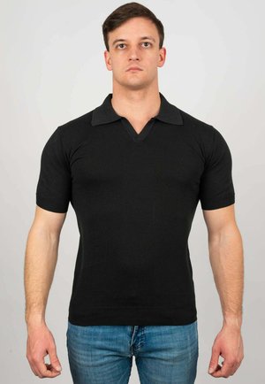 Giovane uomo adulto in piedi di fronte, indossa una polo nera a maniche corte e jeans blu su uno sfondo semplice.