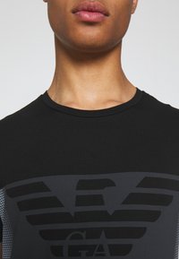 Svart t-shirt med rund halsringning, prytt med en mörkgrå logotyp och randigt mönster. Sidopanel i mesh ger textur och andningsförmåga.